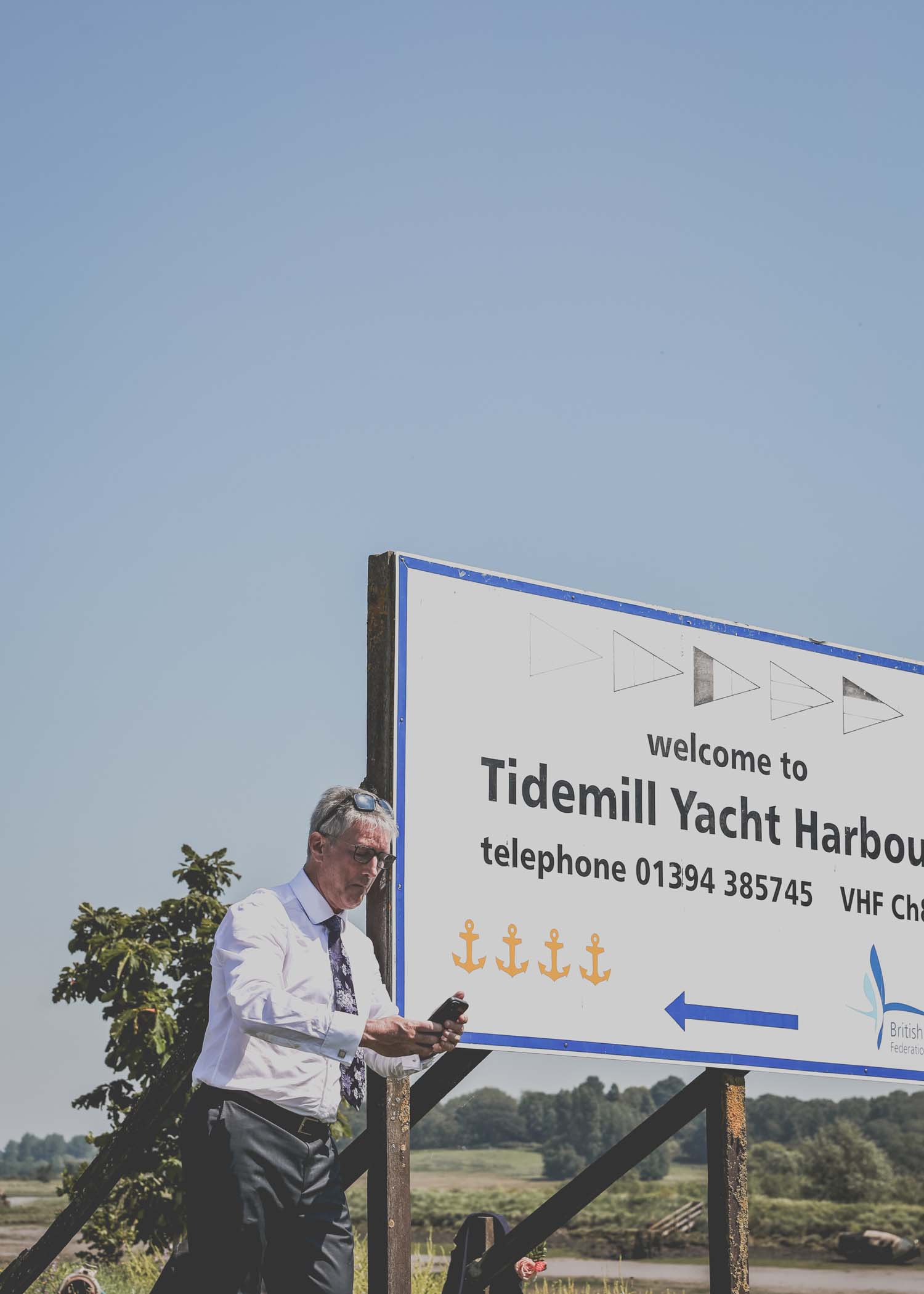 A groom leaning on the harbour sign at woodbridge marina on a wedding day in Woodbridge captured by Suffolk Wedding Photographer Hayley Denston Photography